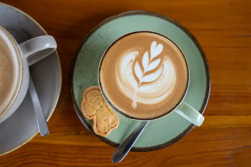 A steaming cup of latte art coffee and a bowl of snacks on a table